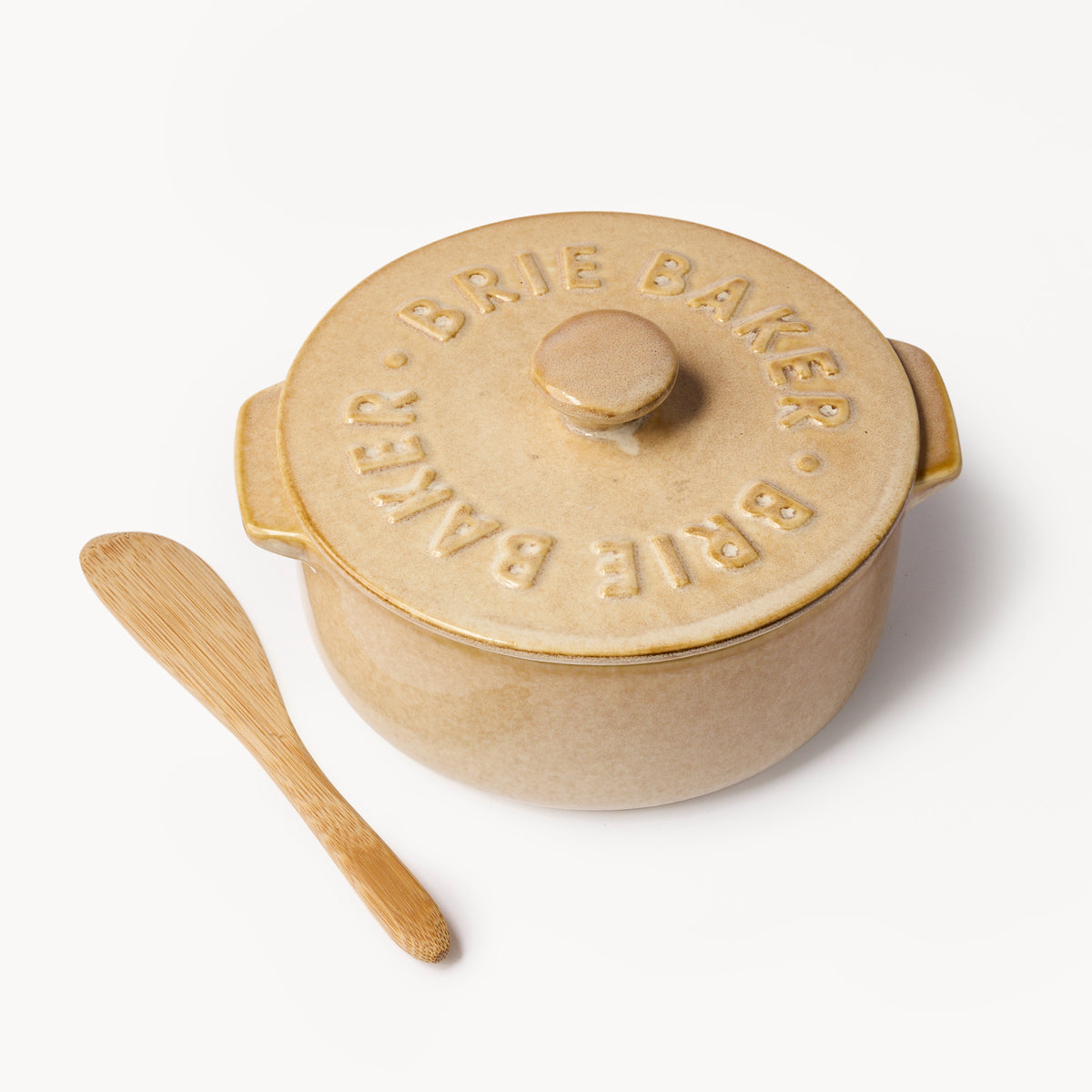 A beige ceramic Brie Baked pot with its lid on, shown next to a small wooden spoon on a clean white background. The lid features embossed text reading 'BRIE BAKER.