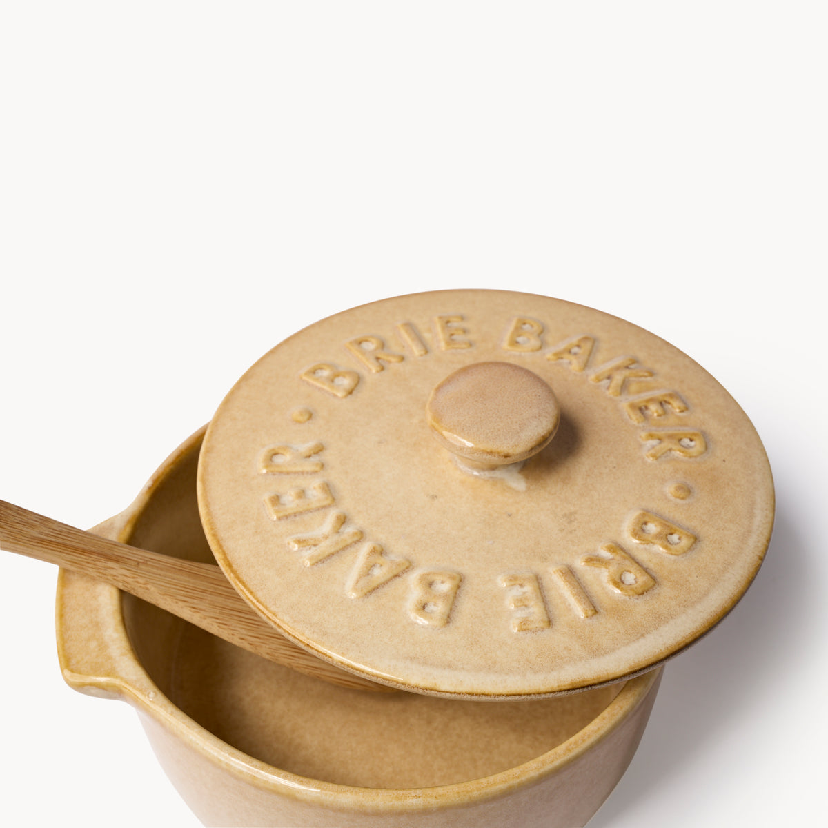 A ceramic Brie Baker with a matching lid embossed with 'BRIE BAKER' and a wooden spoon resting inside, shown on a clean white background.