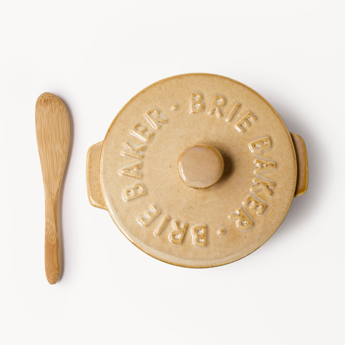 A beige ceramic Brie Bakker lidded pot with a matching wooden spatula, photographed from directly above on a clean white background.