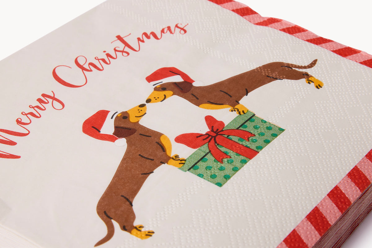 A close-up of a Christmas-themed paper napkin featuring two dachshunds wearing red Santa hats and reaching for a green gift box with a red bow, with the text 'Merry Christmas' in red script. The napkin has a white textured background and red-and-white striped edges.